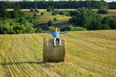 A guy sitting on a large bale of straw in an empty field of cut wheat
