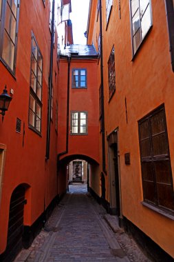 Stockholm 'ün eski kasabasındaki turuncu renkli canlı binalar arasında dar bir geçit var. Duvarları pencerelerle kaplı kemerli bir tünelden geçen kaldırım taşı bir yol var..