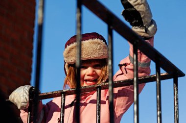 Rahat bir kürk şapkası ve pembe kışlık paltosu olan neşeli bir çocuk, canlı mavi gökyüzüne karşı metal korkuluklardan neşeyle geçiyor. Bu büyüleyici an, çocukluk merakının ve maceranın özünü yakalar. Genç kaşif sınırlarda gezinirken.