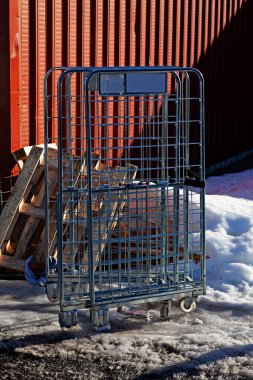 Tek bir metal alışveriş arabası karla kaplı bir araziye ve kırmızı tuğlaya karşı duruyor ve endüstriyel araç ile doğal elementler arasındaki katı zıtlığı yakalıyor. Soğuk, mavi renkli metal çerçeve görsel bir öykü yaratıyor.