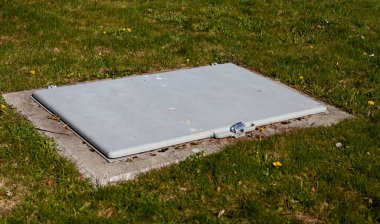 Gri beton bir kiler kapısı ya da bodrum girişi beton bir temele kurulmuş, yemyeşil çimlerle çevrili, etrafa saçılmış küçük sarı karahindibalar..