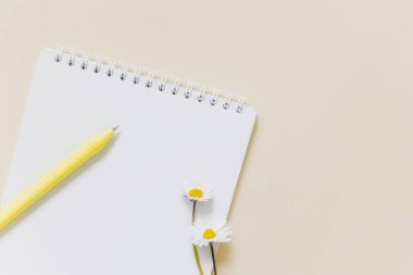 Boş sayfalı defter, papatya çiçekleri ve açık sarı arka planda kalem. Düz yatıyordu. Metin için yer.