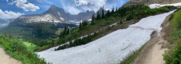 Panorama view of the Iceberg Lake Trail at Glacier National Park, Montana where hikers needed to walk across a narrow path across two glaciers to complete the journey and view a valley of pine trees.