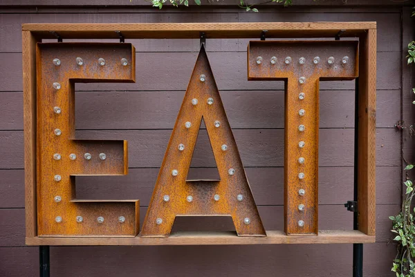 Large, rectangular sign that reads EAT which has lightbulbs and is rusty in color, stands before a wooden plank wall that is painted brown.