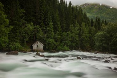 Moskog ve Balestrand arasındaki Norveç manzaralı Gaularfjellet rotası boyunca, bulutlu ve fırtınalı bir günde bir dere akıntısı. Yanlız bir ahşap ev kenarda duruyor.. 