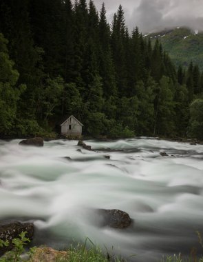 Moskog ve Balestrand arasındaki Norveç manzaralı Gaularfjellet rotası boyunca, bulutlu ve fırtınalı bir günde bir dere akıntısı. Yanlız bir ahşap ev kenarda duruyor.. 