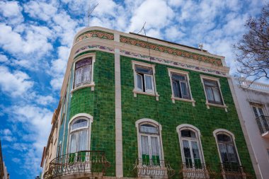 An apartment building with green tiles on it's facade, photographed in Lagos, Portugal during a sunny afternoon with clouds in the sky.  clipart