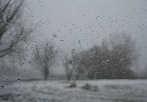 wet window background, raindrops on glass, silhouettes of trees against a gray sky, a snow-covered beach near a frozen river