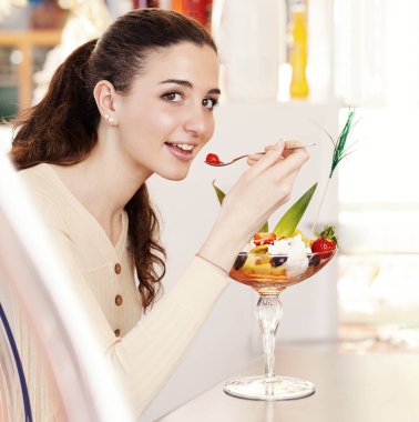 Kafe kafeteryasında, dondurmacıda ve pastanede üzerinde dondurma olan meyve salatasıyla gülümseyen güzel bir genç kadın. Dondurma yiyen esmer kızın portresi, kirazlı meyve..