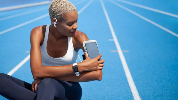 Smiling athletic sportswoman wears earphones and touch smartphone in her armband. Tracks her running sports training and jogging. African American woman sitting on the Olympic track with blue lanes