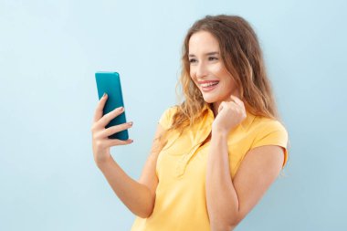 Neşeli ve güzel bir genç kadın gülerken cep telefonuna bak, rahat giyin, internet tarayıcı sayfasında sörf yap ya da akıllı telefon uygulaması kullan. Çevrimiçi yaşam tarzı konsepti. Açık mavi arkaplanda izole