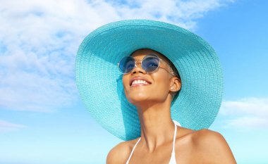 Kumsalın kenarında turkuaz güneş şapkası, mavi güneş gözlüğü ve bikini takan mutlu genç bir kadın. Güneşli bir yaz gününde mavi gökyüzü ile Afrika kökenli Latin Amerikalı bir kadının portresi.