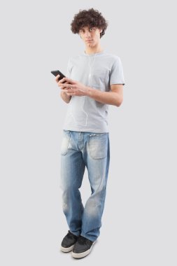 Genç bir adam, gülümseyen ve yakışıklı, kulaklıklı akıllı telefonunu müzik dinlemek ya da sesli mesaj dinlemek için kullanıyor. Mavi gözleriyle kameraya bakıyor. Gri arka planda tüm vücudunu çekiyor.