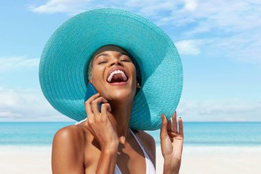 Kumsalda mavi güneş şapkası takan ve güneşli bir günde mavi gökyüzü olan cep telefonu kullanan mutlu bir kadın. Yaz tatili kavramı, online alışveriş, seyahat ve tatil yeri rezervasyonu