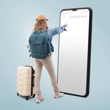 Sırt çantası ve tramvay çantasıyla seyahat eden genç bir turist akıllı telefon ekranını işaret ediyor. Mavi arka planda izole edilmiş. Tatil kavramı veya online rezervasyon