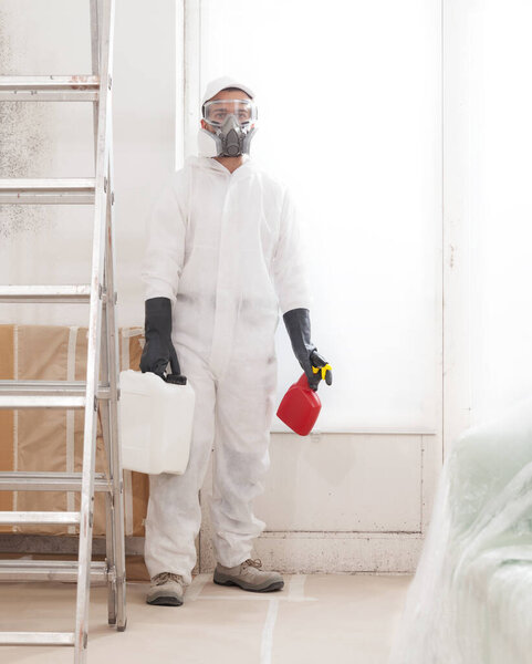 Professional mold removal service: Worker wearing protective suit, gloves and respirator mask, spraying product to clean moldy wall before room painting work. Home refurbishment site with window