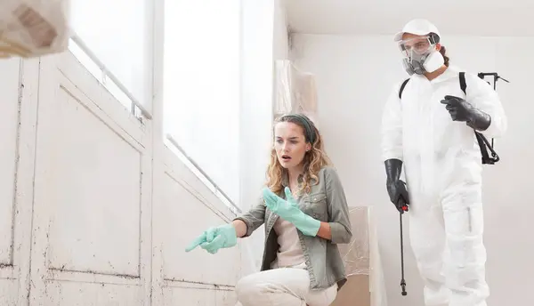stock image Mold remediation service: worried woman at a home renovation site shows a mold problem to a professional worker clad in a protective suit and a backpack sprayer to remove mold before house painting