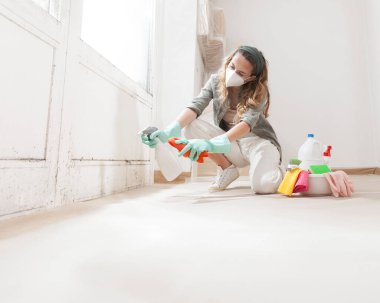 Evin duvarındaki küfleri sprey şişeyle temizleyen kadın, eldiven takıyor ve koruyucu bir maske takıyor. Evi yenilemeden ve boyamadan önce küfü düzeltmek için ev temizlik ürünleri kullanılıyor.