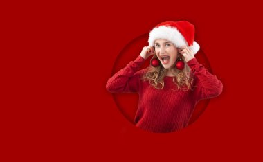 Noel Baba şapkası takmış neşeli sarışın bir kadın, Noel toplarıyla oynuyor sahte küpeler takıyor, kırmızı arka planda izole edilmiş. Şablon Satılık ve promosyonlar ya da çevrimiçi alışveriş reklamları sunmak