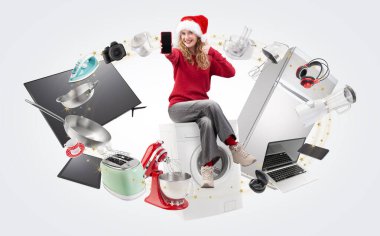 Electronics and appliance store Christmas shopping, smiling woman showing cellphone and thumb up sitting on washing machine with household appliances, top offers and sales of Christmas shopping online clipart