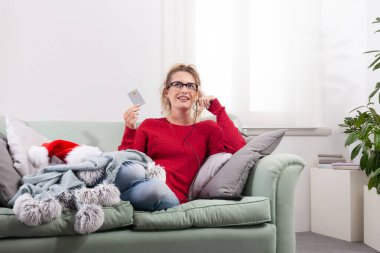 Bir kadın evdeki kanepesinde rahat bir şekilde oturuyor, akıllı gözlüklerini Noel alışverişi için internette gezinirken, kredi kartını akıllı telefonuyla satın alma ve ödeme yapmak için kullanıyor.