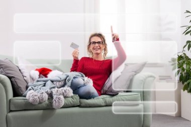 Bir kadın evdeki kanepesinde rahat bir şekilde oturuyor, akıllı gözlüklerini Noel alışverişi için internette gezinirken, kredi kartını akıllı telefonuyla satın alma ve ödeme yapmak için kullanıyor.