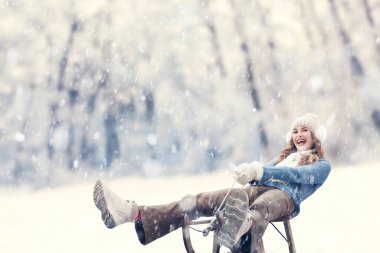 Happy young woman in snowfall sledding and enjoying the ride on sled outdoor, dressed in warm winter clothing, hat, scarf, and gloves. A ski trip during the Christmas holidays in the winter season clipart