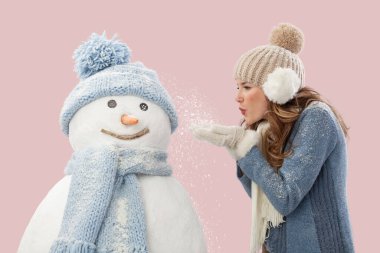 Mutlu genç bir kadın pembe arka planda izole edilmiş bir kardan adama kar üflüyor. Kış mevsiminde Noel tatilinin tadını çıkarıyorum. Soğutma ve ısıtma sıcaklığı kavramı