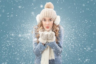 Mavi arka planda kar yağışı altında mahsur kalmış mutlu kadın kameraya bakarak ellerinden kar üflüyor. Noel tatili ya da kış tatili ve soğutma ve ısıtma sıcaklığı kavramı