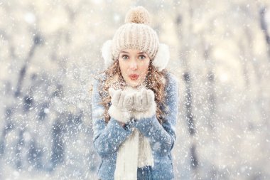 Kar yağarken, dışarıda kameraya bakarak ellerinden kar üfleyen mutlu genç bir kadın. Noel tatili ya da kış tatili ve soğutma ve ısıtma sıcaklığı kavramı