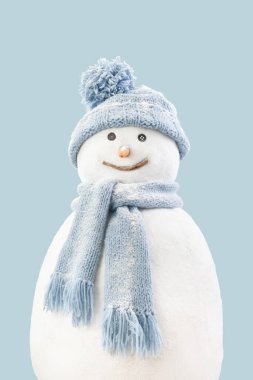 Mavi arka planda izole bir şekilde kameraya bakan örgü örülü kış eşarbı ve şapka giyen mutlu gerçekçi kardan adam. Noel tatili ya da kış tatili ve soğutma ve ısıtma sıcaklığı kavramı
