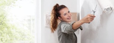 A happy, smiling young woman with a paint roller painting white wall near a bright window overlooking a green panorama at home. She is setting up her new place after moving out. Green home renovation clipart