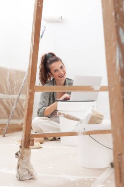 Ev tadilatı sırasında dizüstü bilgisayarını kullanarak duvar boyası arayan ve alan mutlu bir kadın. Bir boya kovası ve ahşap merdivenin yanında oturmuş internetten boya dükkanından alışveriş yapıyor.