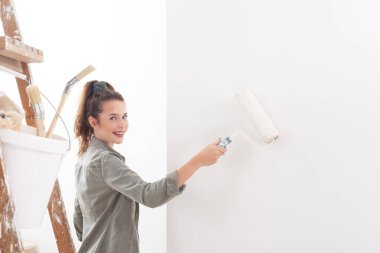 Mutlu gülümseyen genç bir kadın evde boyacı silindiri ve boya fırçalarıyla beyaz bir duvar boyuyor. Taşındıktan sonra yeni evini kuruyor. Satın alma ve ev yenileme konsepti