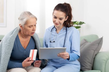 Genç bir hemşire ya da doktor, yaşlı bir kadına dijital tabletteki ilaçları açıklarken ve gösterirken gülümsüyor. Evde, bir klinikte ya da bakımevinde. Sağlık hizmetleri kavramı