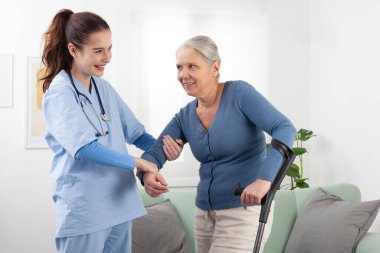 Koltuk değnekleriyle yürümesine yardım ederken yaşlı bir kadınla ilgilenen hemşire ya da doktor. Klinikte ya da hastanede evde bakım. Sağlık ve sigorta kavramı