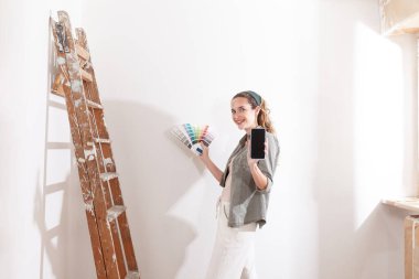 Boya örnekleri ve telefon gösteren mutlu bir kadın. Duvarları boyamak için bir renk seçin. DIY, taşındıktan sonra evi dekore etmek, yenilemek ve yenilemek için çalışır.