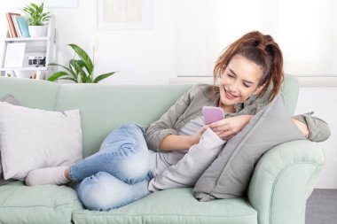 Rahat rahat gülümseyen genç bir kadın rahat bir kanepede rahat bir kıyafetle uzanıyor, elinde bir akıllı telefon ve mesajlar, sosyal medya ve dijital içerik tutuyor.