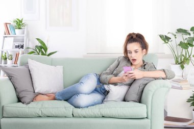 Rahat genç bir kadın rahat bir kanepede rahat bir kıyafetle uzanmış elinde akıllı bir telefonla mesajları, sosyal medyayı ve dijital içeriği karıştırıyor.