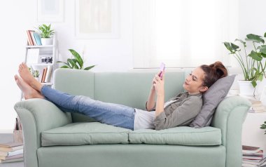 Rahat rahat gülümseyen genç bir kadın rahat bir kanepede rahat bir kıyafetle uzanıyor, elinde bir akıllı telefon ve mesajlar, sosyal medya ve dijital içerik tutuyor.