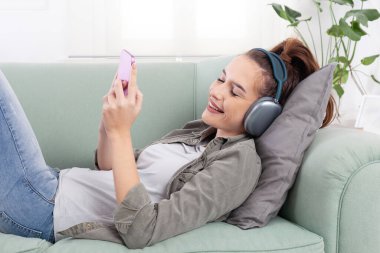 Genç, rahat ve gülümseyen bir kadın rahat bir kanepede uzanmış, elinde akıllı bir telefonla mesajları, sosyal medyayı, müziği ve dijital içeriği inceliyor. Kulaklık takıyor, modern, bağlı bir yaşam tarzı var.