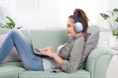 Genç, rahat ve gülümseyen bir kadın rahat bir kanepede uzanıyor, mesajları, sosyal medyayı ve dijital içeriği dinlemek için bilgisayarını kullanıyor, kulaklıklarıyla müzik dinliyor, modern, bağlı bir yaşam tarzı.