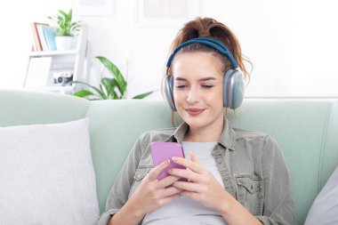 Genç, rahat ve gülümseyen bir kadın bir koltukta oturmuş, elinde akıllı bir telefonla mesajları, sosyal medyayı, müziği ve dijital içeriği inceliyor. Kulaklık takıyor, modern, bağlı bir yaşam tarzı var.