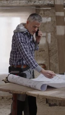 Bir konut yenileme sahasında inşaat işçisi proje bina özelliklerini doğrulamak için telefonda konuşurken ayrıntılı ev planları ve planlarını gözden geçirir..