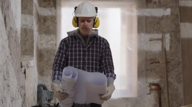 Kafası karışmış inşaat işçisi, bir yenileme şantiyesinde çalışıyor. Evin planlarını ve planlarını okuyor. Kafası karışmış bir şekilde etrafa bakıyor ve proje planını, görevlerini ve çalışmalarını anlamaya çalışıyor.