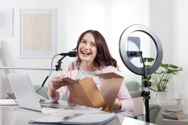 Gülümseyen kadın etkiler akıllı telefon ve halka ışığı ile evden canlı yayında, sponsorluk veya tasarımcı markası için boks ve ürün değerlendirmesini kaydediyor, seyirci ve takipçilerle çevrimiçi paylaşıyor