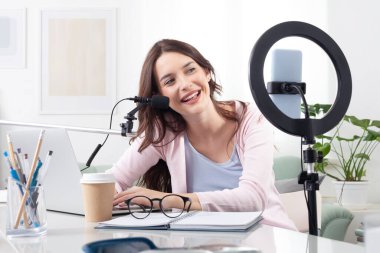 Gülümseyen genç bayan içerik yaratıcısı ve sözü geçen, akıllı telefon ve halka ışığı kullanan, dizüstü bilgisayar ve mikrofonla çalışan. Takipçiler için canlı yayın veya kayıt videoları veya dijital içerik