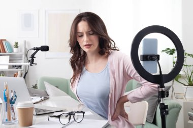 Yorgun kadın içerik yaratıcısı ve sözü geçen kişi, ağrısını masasında akıllı telefon ve halka ışığıyla geriyor. Video dersleri, podcastlar ya da sosyal medya içeriği oluşturulmasından çok çalışılmış