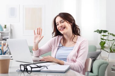 Evdeki mutlu genç kadın, masada oturuyor ve dizüstü bilgisayarına el sallarken gülümsüyor. Olumlu bir uzaktan çalışma ya da çevrimiçi öğrenme, video çağrısı, neşeli bir ev ortamında dijital işbirliği temsil etmek