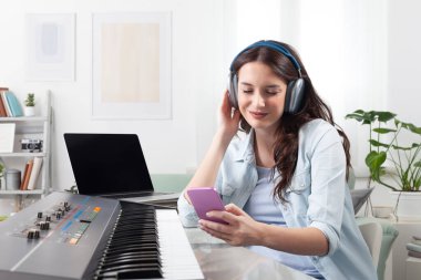 Evde müzik klavyesi çalan, kulaklıkla dinleyen ve akıllı telefonuyla gezinen genç bir kadın. Müzik öğrenmek ve bestelemek. Yaratıcı iş kavramı, müzikle dolu bir yaşam tarzı hayal etmek.